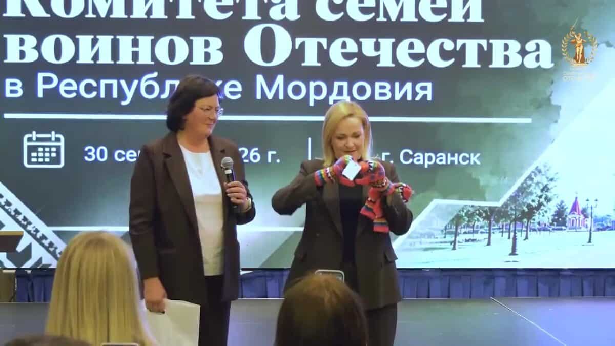 Две женщины на сцене демонстрируют полосатое вязаное изделие перед аудиторией.