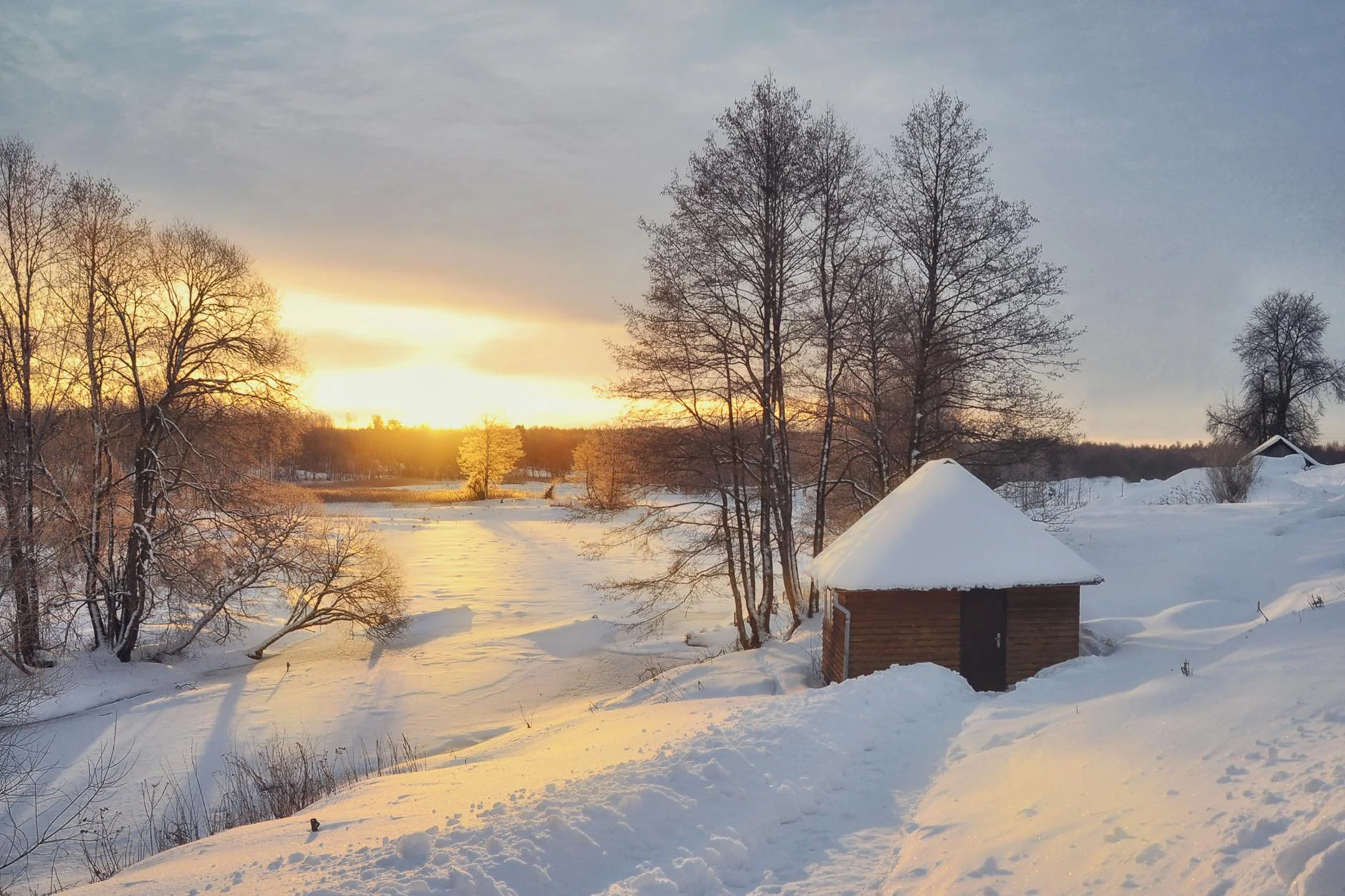 Scenic winter sunrise over snow-covered cabin and trees by a frozen lake.